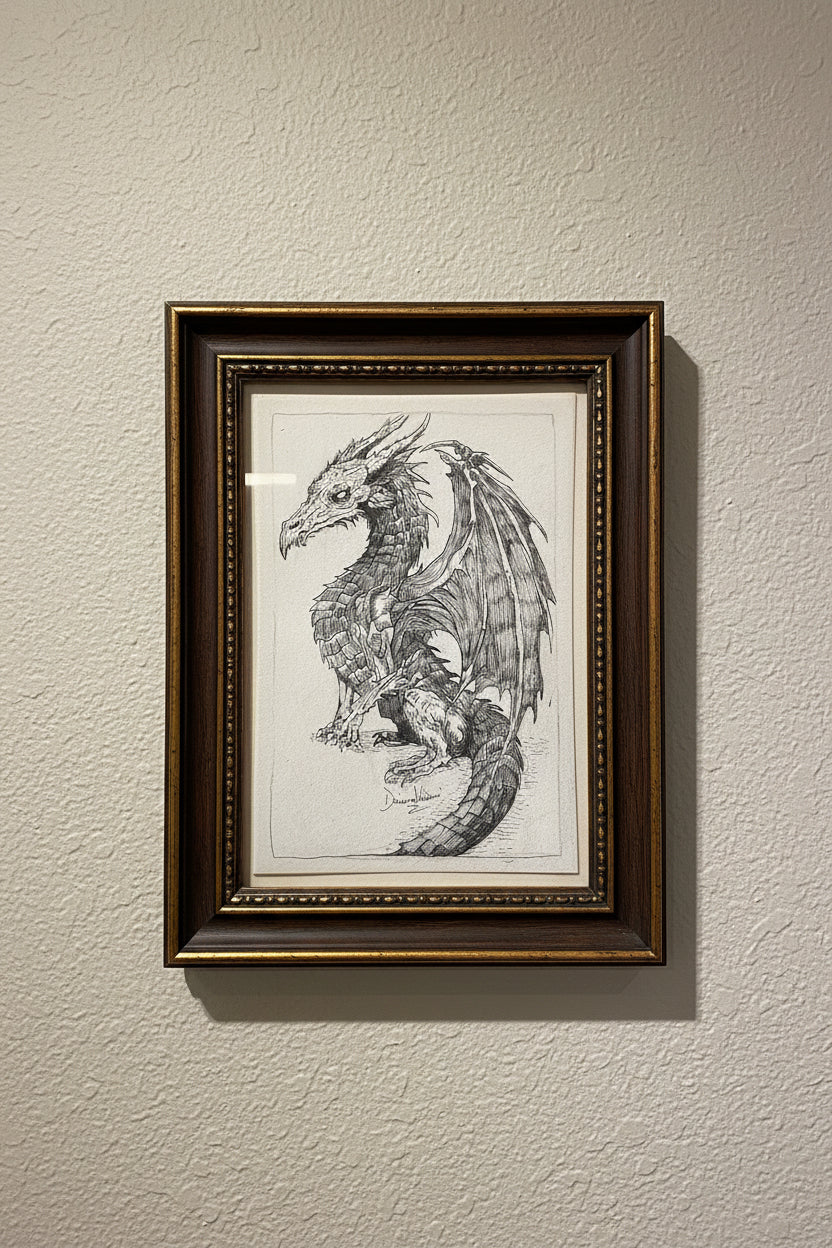 A detailed black and white hand-drawn sketch of a fantasy dragon on a spiral-bound sketchbook page, photographed in a desk setting.