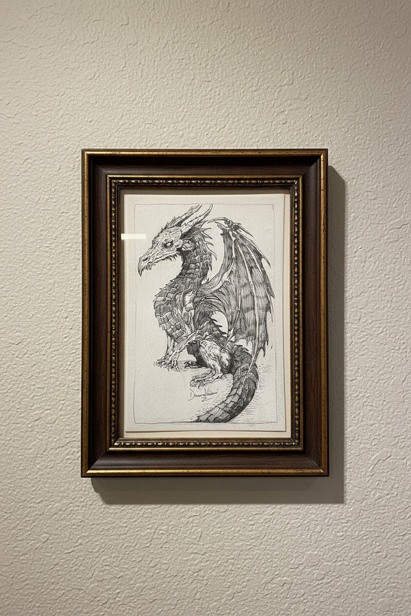 A detailed black and white hand-drawn sketch of a fantasy dragon on a spiral-bound sketchbook page, photographed in a desk setting.